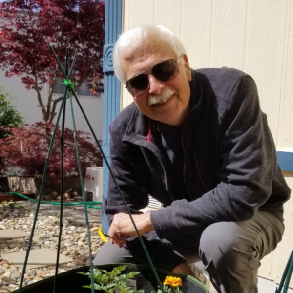 Mike Robinson kneels by a plant in a garden. He is smiling and wearing sunglasses on a sunny day.