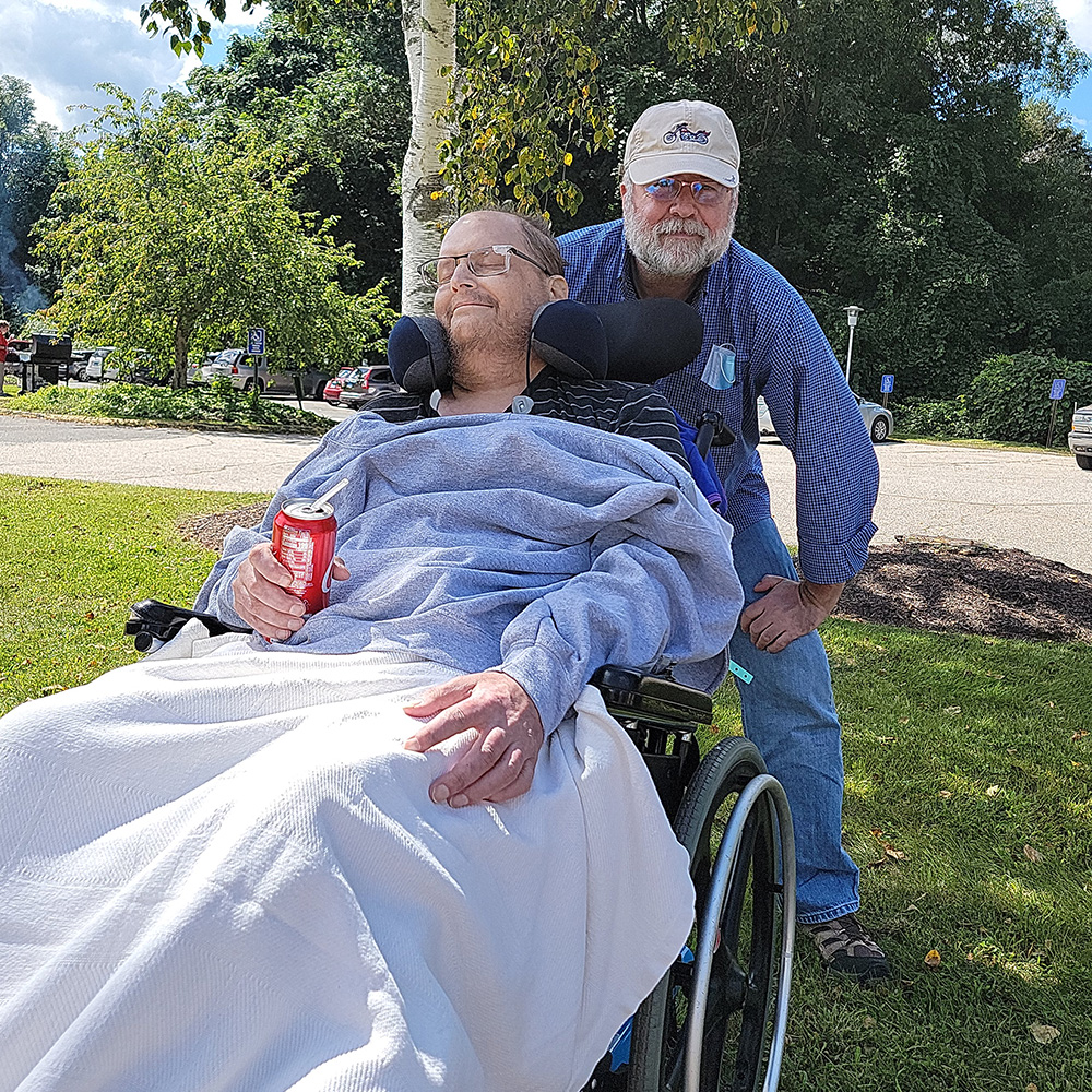 Clifford Biniarz in his wheelchair outdoors holding a coke with his brother Tyrone behind him.